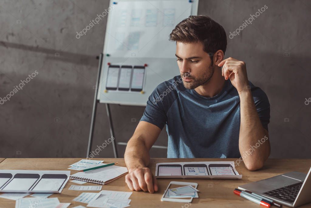 Selective focus of handsome designer planning mobile app with layouts on table stock vector