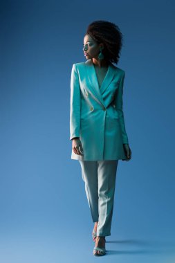  Çekici Afro-Amerikalı takım elbiseli kadın mavi arka plana bakıyor. 