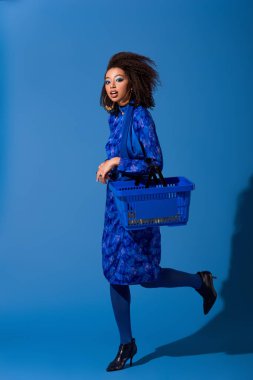 Afro-Amerikalı bir kadın mavi arka planda alışveriş sepeti tutuyor. 