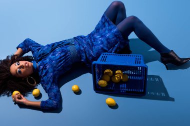 Alış veriş sepeti ve mavi arka planda limonlarla yatan Afrikalı Amerikalı kadın. 