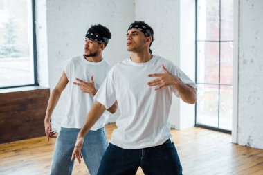 Saç bantlı yakışıklı çok kültürlü dansçılar hip-hop dansı yaparken el kol hareketi yapıyor ve poz veriyorlar.