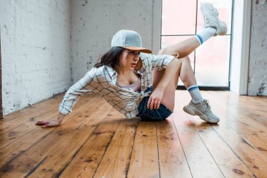 beautiful dancer in cap sitting on floor and dancing hip-hop in dance studio 
