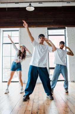 handsome multicultural dancers in headbands and attractive girl in cap standing with outstretched hands 