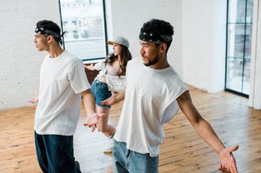 selective focus of multicultural men in headbands and young woman in cap dancing hip-hop 