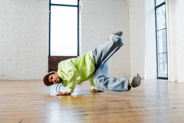 trendy african american man in cap breakdancing in dance studio 