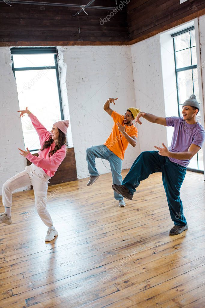 attractive girl breakdancing and gesturing with stylish multicultural men in hats 