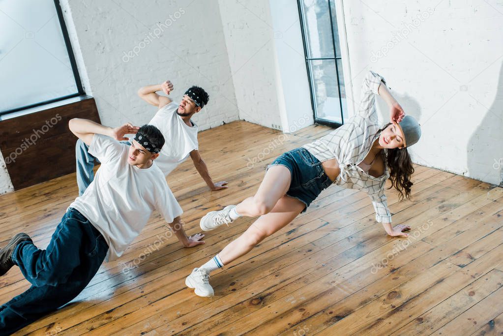 trendy girl touching cap while breakdancing with multicultural dancers in dance studio 