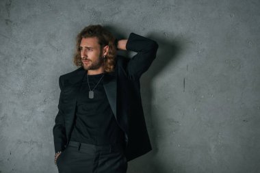 Siyah takım elbiseli şık bir işadamı. El ele poz veriyor ve gri duvarın yanındaki koyu renkli duvara bakıyor.