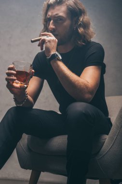 Siyah kıyafetli, puro içen ve gri duvarın yanındaki koltukta viski içen bir iş adamı.