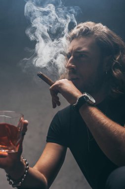 Gri duvarın yanında puro içip viski içen siyah kıyafetli gergin bir işadamı.