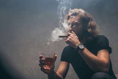 Siyah kıyafetli, puro içen ve gri duvarın yanında viski içen bir iş adamı.