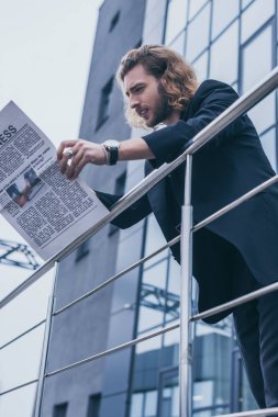 Siyah takım elbiseli moda işadamının ofis binası ve korkuluk yakınında gazete okumasının düşük açılı görüntüsü