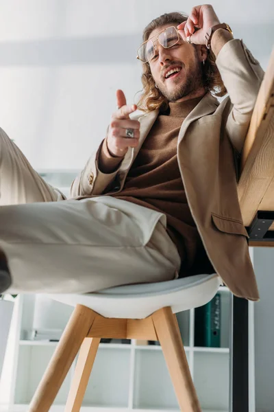 坐在办公室椅子上的时髦商人一边微笑着一边用手指指着相机 — 图库照片