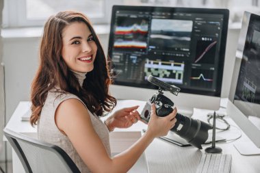 Neşeli sanat editörü bilgisayar monitörlü dijital kamerayı masanın yanında tutuyor 