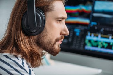 Bilgisayar monitörünün yanındaki kulaklıkla müzik dinleyen sakallı editörün yan görüntüsü