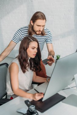 Sakallı sanat editörü çekici bir kadının yanındaki bilgisayar monitörünü işaret ediyor. 