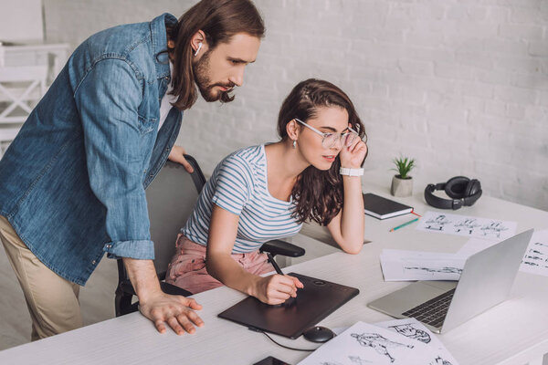 illustrator looking at laptop near coworker in glasses and sketches 