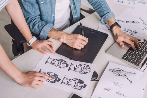 cropped view of illustrator pointing with finger at cartoon sketch near coworker 