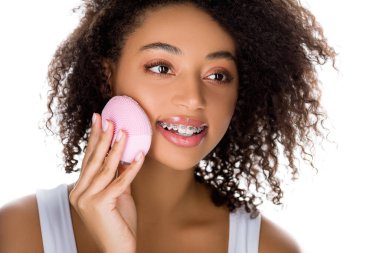 attractive happy african american girl with braces using silicone cleansing facial brush, isolated on white clipart