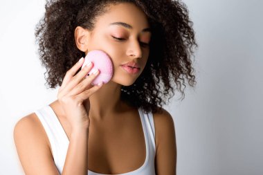 Kıvırcık Afrikalı Amerikalı kız silikon temizleyici yüz fırçası kullanıyor, gri renkte izole edilmiş.