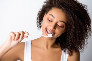 Diş telleriyle diş fırçasına bakan neşeli Afro-Amerikalı kadın, gri renkte izole edilmiş.