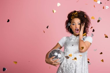 shocked african american girl in paillettes dress holding disco ball, isolated on pink with confetti clipart