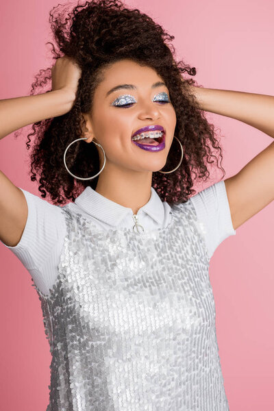 positive african american girl with dental braces, with silver glitter eyeshadows and purple lips wearing paillettes dress, isolated on pink