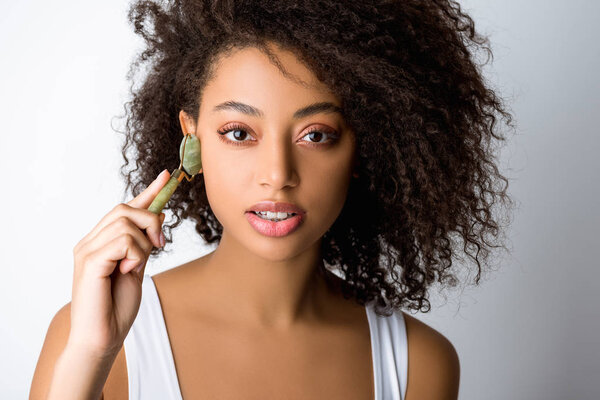 attractive african american girl using stone facial roller, isolated on grey 