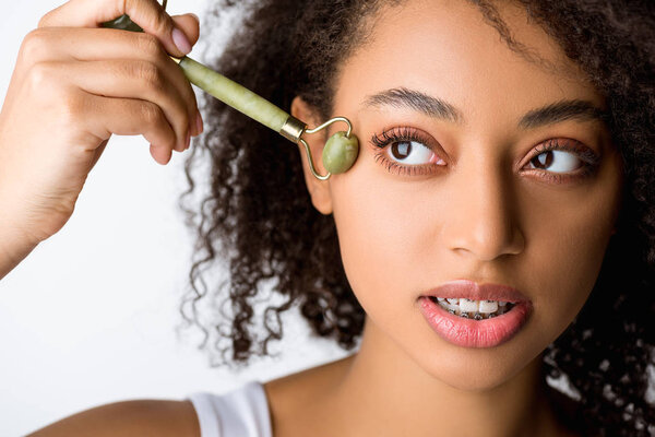 beautiful curly african american girl using jade facial roller, isolated on grey 