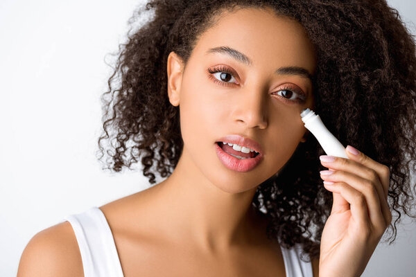 attractive curly african american girl using moisturizing eye roller, isolated on grey