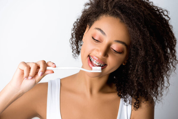 cheerful african american woman with dental braces looking at toothbrush, isolated on grey