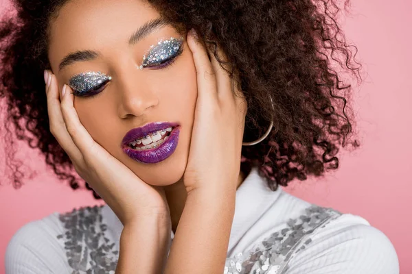 tender african american woman with silver glitter eyeshadows and purple lips wearing paillettes dress, isolated on pink