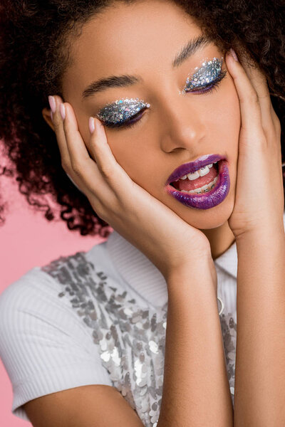 emotional african american girl with silver glitter eyeshadows and purple lips wearing paillettes dress, isolated on pink
