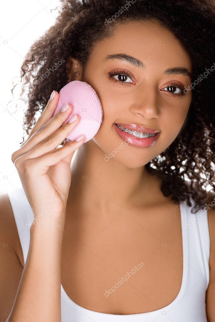 beautiful smiling african american girl with orthodontic cases using silicone cleansing facial brush, isolated on white