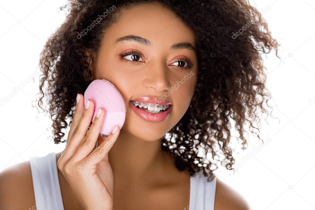 attractive happy african american girl with braces using silicone cleansing facial brush, isolated on white