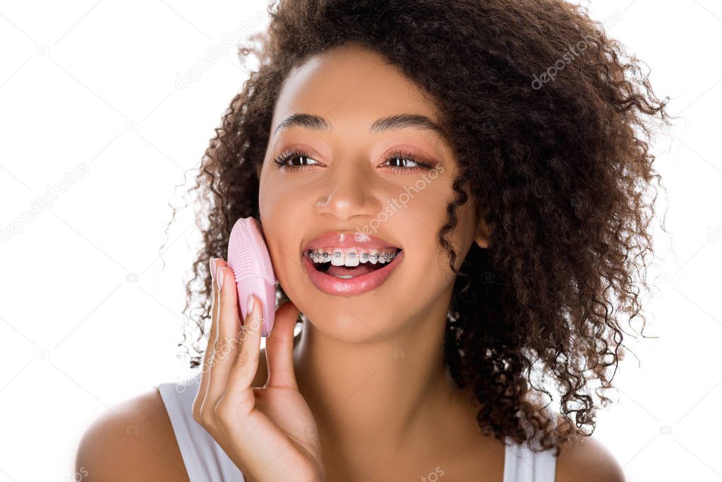 smiling african american girl with dental braces using silicone cleansing facial brush, isolated on white
