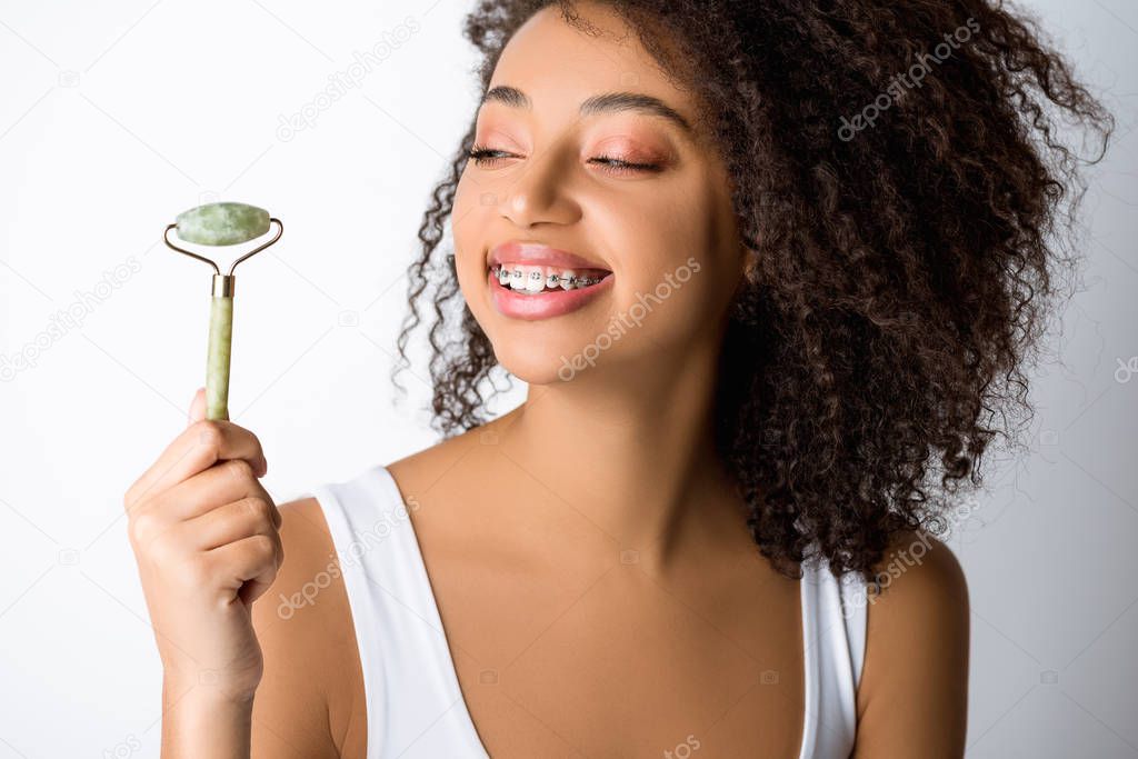 happy african american girl with dental braces using natural massage roller, isolated on grey 