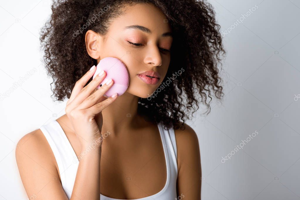 curly african american girl using silicone cleansing facial brush, isolated on grey