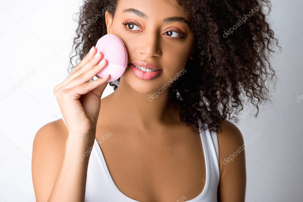 african american girl using silicone cleansing facial brush, isolated on grey