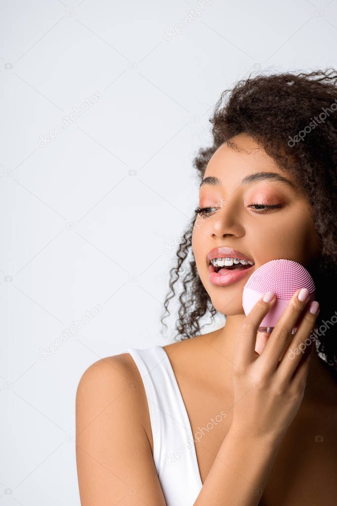 smiling african american woman with orthodontic cases using silicone cleansing facial brush, isolated on grey