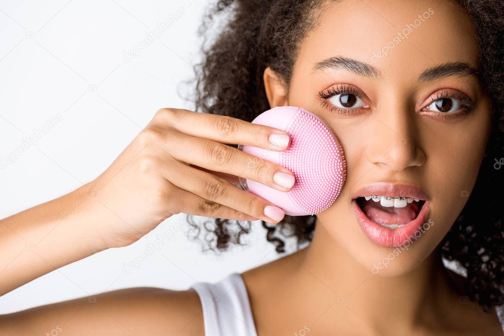 surprised african american girl with dental braces using silicone cleansing facial brush, isolated on grey