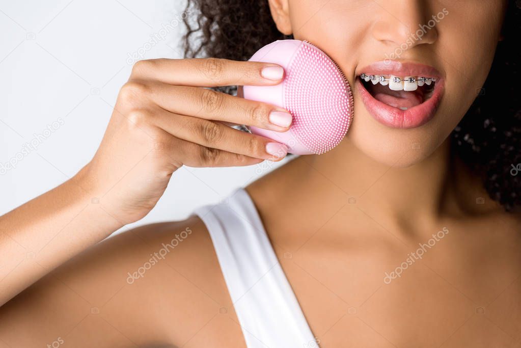 cropped view of surprised african american girl with dental braces using silicone cleansing facial brush, isolated on grey