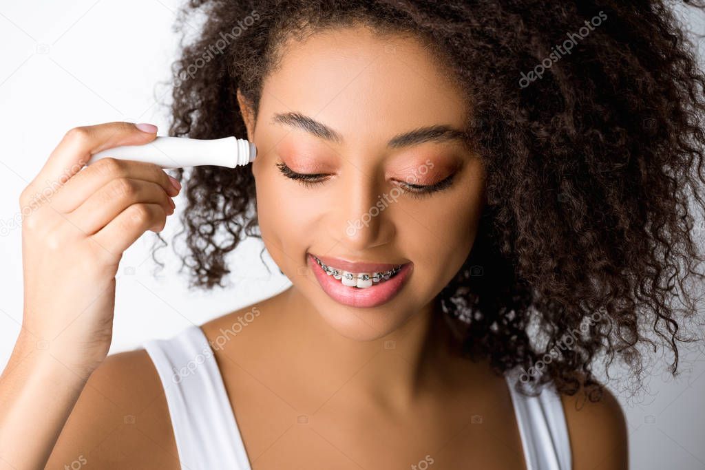 smiling african american girl with dental braces using moisturizing eye roller, isolated on grey