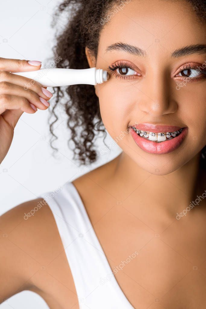 happy african american woman with dental braces using moisturizing eye roller, isolated on grey