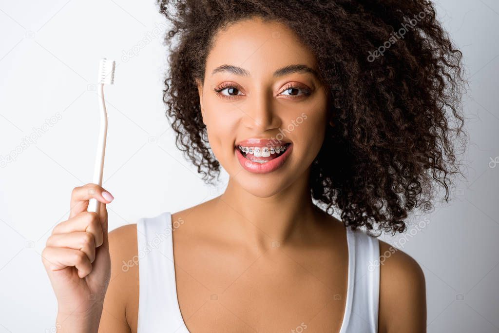 happy african american woman with dental braces holding toothbrush, isolated on grey