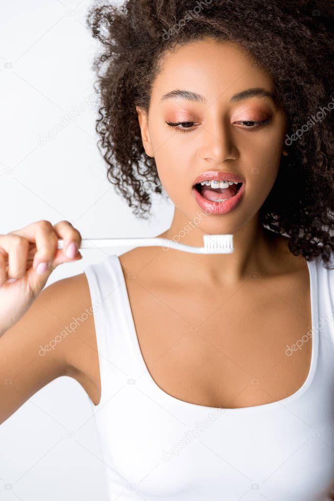 shocked african american woman with dental braces looking at toothbrush, isolated on grey