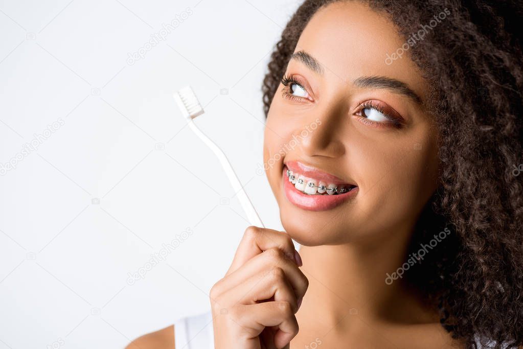 beautiful curly african american woman with dental braces looking at toothbrush, isolated on grey