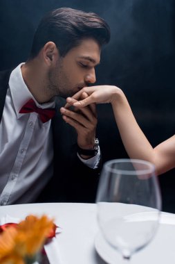 Siyah arka planda servis masasında oturan, takım elbiseli bir adamın el öptüğü, dumanlı bir kadın.