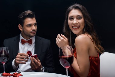 Smiling woman looking away by handsome man with jewelry ring at served table isolated on black clipart
