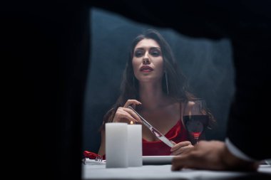 Seçici odak noktası, elinde bıçak olan güzel bir kız ve siyah arka planda romantik bir akşam yemeği sırasında erkek arkadaşına bakan duman.